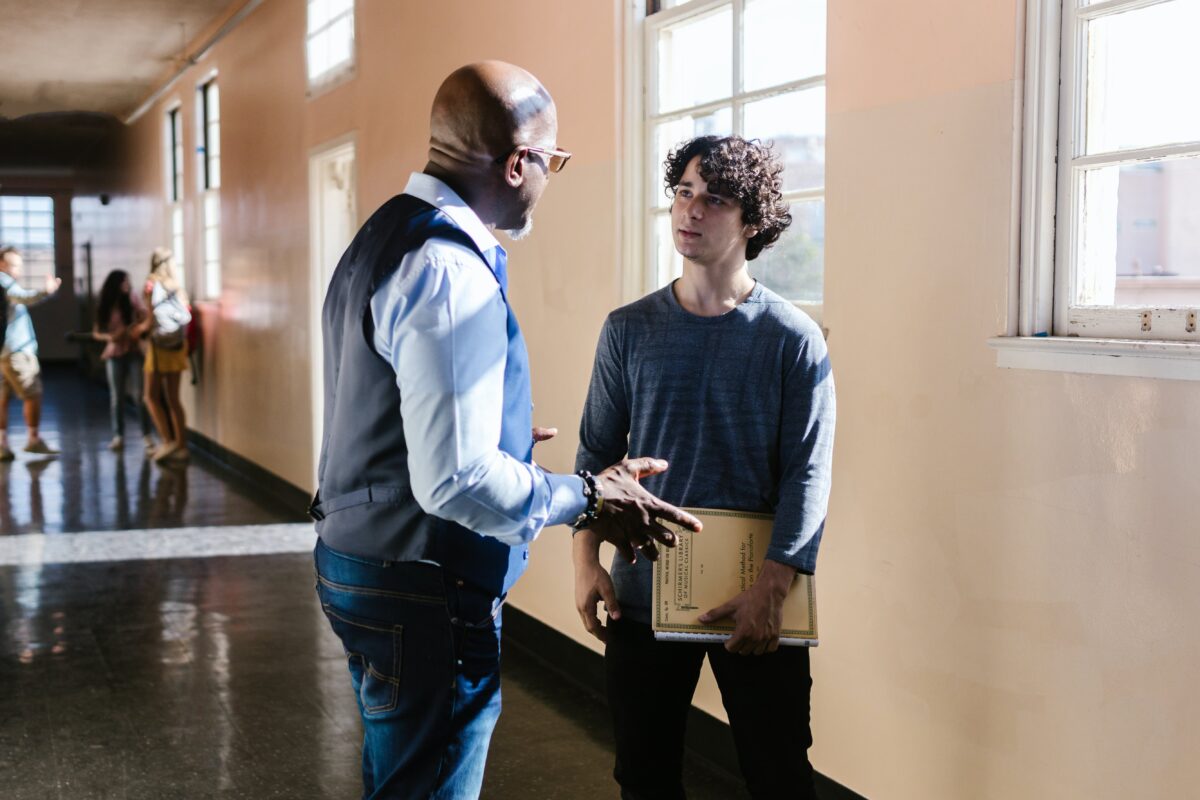 Bald, black male teacher wearing a vest speaking to a white, male student with curly hair holding a notebook.