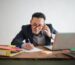 Asian mearing red glasses seated at a desk. He is taking notes on paper, talking on the phone, and looking at a laptop.