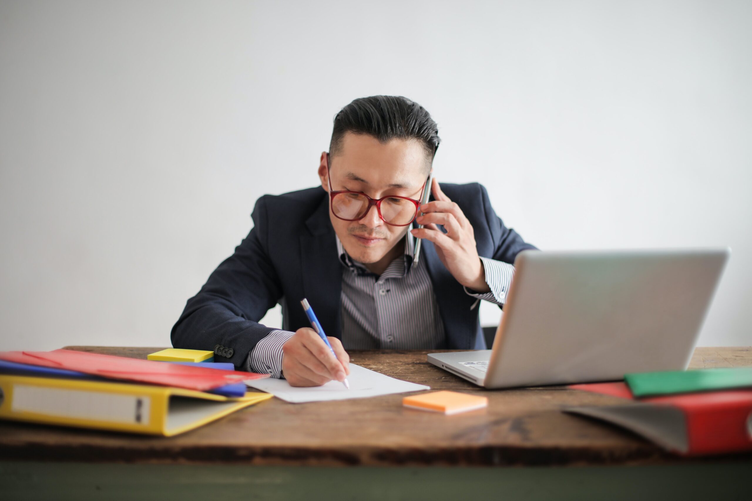 Asian mearing red glasses seated at a desk. He is taking notes on paper, talking on the phone, and looking at a laptop.